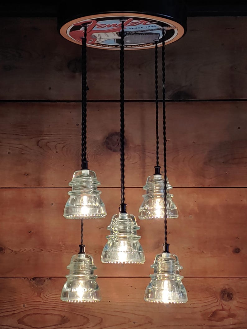 May include: A distinctive light fitting with six clear glass insulator pendant lights hanging from a circular fixture. The fixture has a vintage look, with black cords and a wooden background. The top fixture has the text "Fender Since 1946".