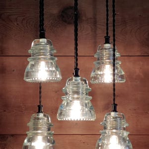 May include: Five pendant lights with clear glass shades, each with a visible light bulb. The shades are shaped like insulators and hang from black cords against a wooden background. The lights are turned on.
