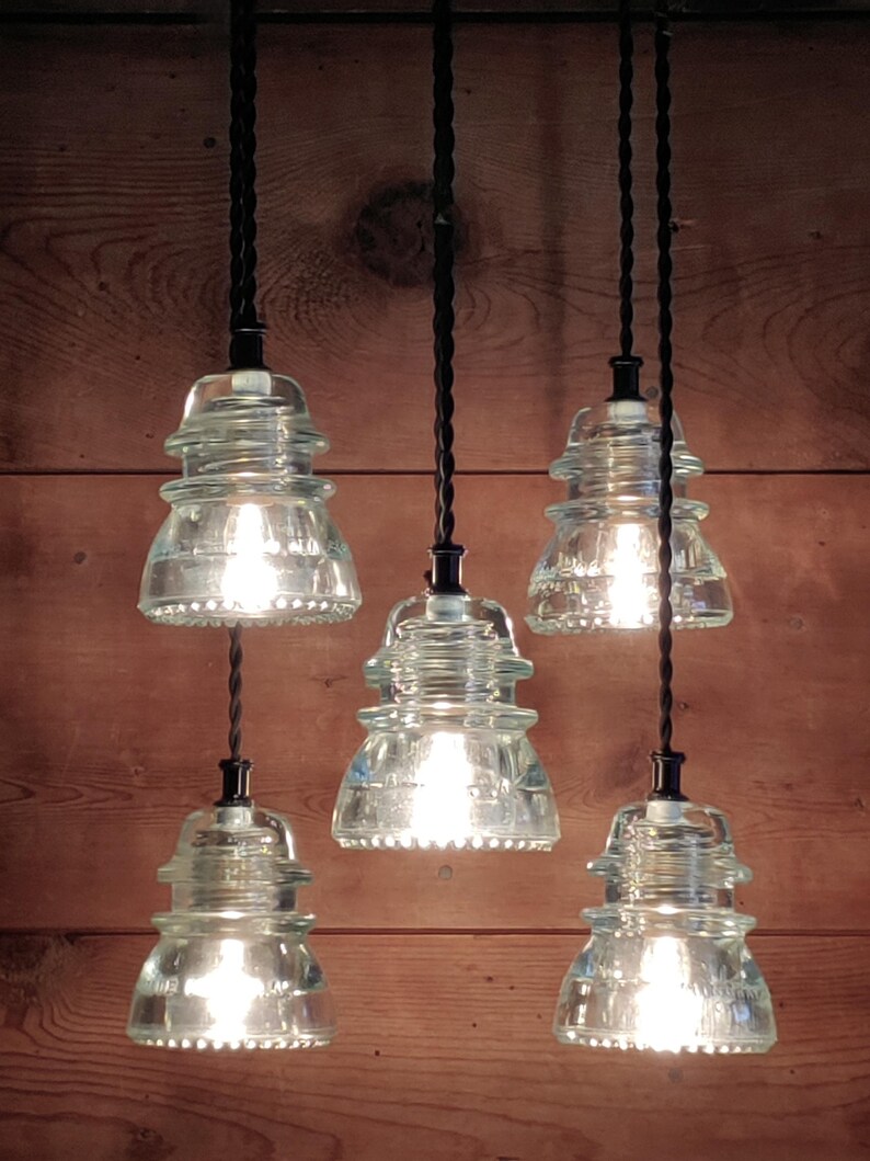 May include: Five pendant lights with clear glass shades, each with a visible light bulb. The shades are shaped like insulators and hang from black cords against a wooden background. The lights are turned on.