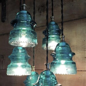 May include: A collection of teal glass insulator pendant lights. The vintage-style light fittings are suspended from black cords, and feature visible text. The exposed bulbs emit a warm light. The lights are a unique, industrial design.