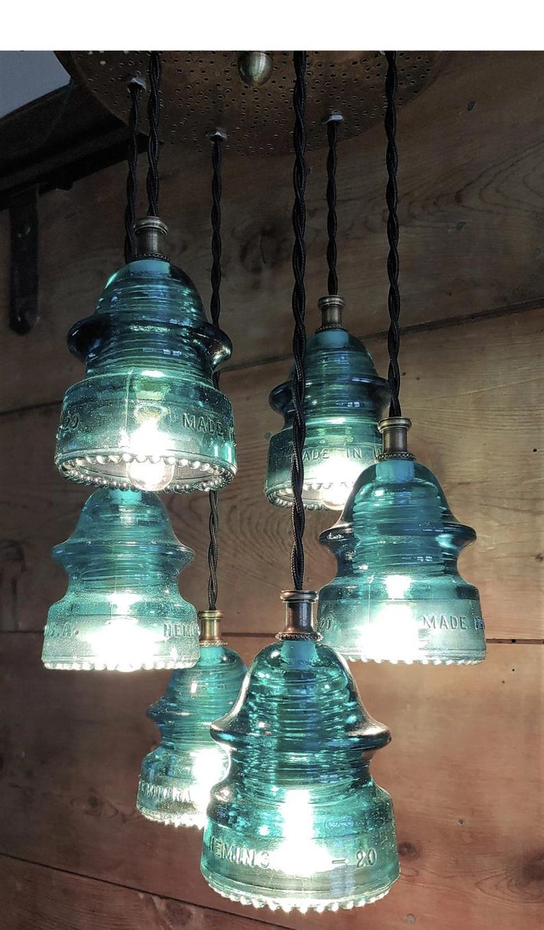 May include: A collection of teal glass insulator pendant lights. The vintage-style light fittings are suspended from black cords, and feature visible text. The exposed bulbs emit a warm light. The lights are a unique, industrial design.