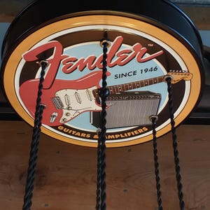 May include: A circular sign with the Fender logo, a red electric guitar, and an amplifier. The sign has a brown background with a light blue centre and the text "SINCE 1946" and "GUITARS & AMPLIFIERS". Black cords hang from the sign.