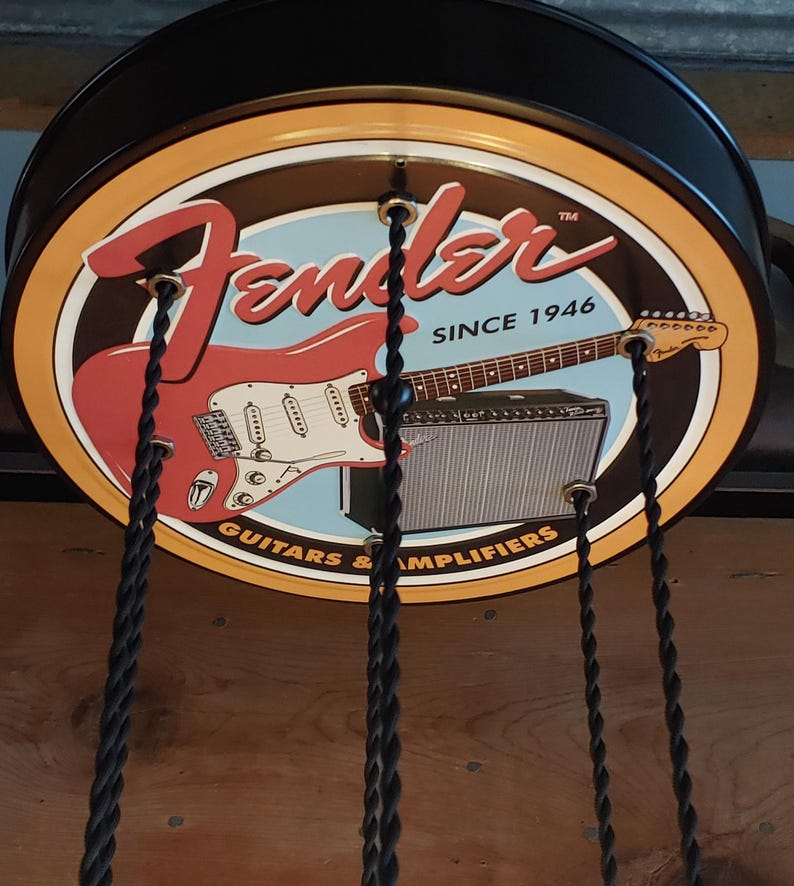May include: A circular sign with the Fender logo, a red electric guitar, and an amplifier. The sign has a brown background with a light blue centre and the text "SINCE 1946" and "GUITARS & AMPLIFIERS". Black cords hang from the sign.
