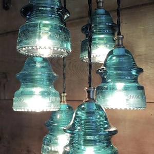 May include: A cluster of teal glass pendant lights with Edison bulbs. Each light is made from a vintage glass insulator, with the words "MADE IN" and other text visible. The lights hang from black cords.