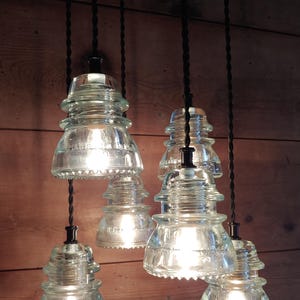 May include: Clear glass pendant lights with an unusual insulator design. Each light hangs from a black cord and has a vintage-inspired shape. The lights are illuminated, casting a warm glow. The background is a wooden wall.