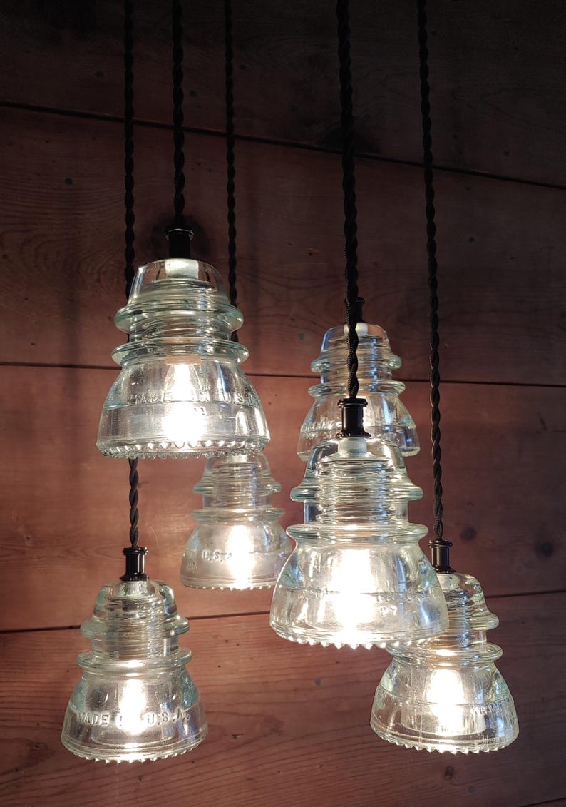 May include: Clear glass pendant lights with an unusual insulator design. Each light hangs from a black cord and has a vintage-inspired shape. The lights are illuminated, casting a warm glow. The background is a wooden wall.