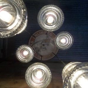 May include: A unique chandelier made from repurposed glass bottles. The clear glass bottles are arranged in a circular pattern, each housing a bright light bulb. The fixture is suspended from the ceiling, creating a warm and inviting ambiance. The design includes a vintage sign.