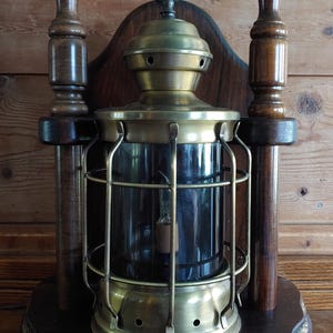 May include: A nautical-style wall lamp featuring a brass lantern with a protective metal cage, mounted on a dark wood backboard. The lamp is flanked by two vertical wooden posts and sits on a circular wooden base accented with a thick rope trim.