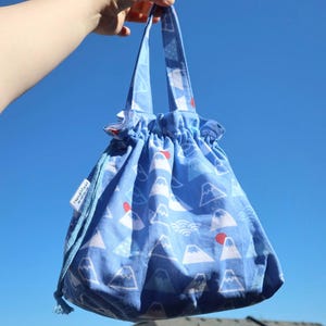 May include: A blue drawstring bag featuring a mountain and sun design. The bag is held up against a clear blue sky. The bag has a white tag that says "handmade by..."
