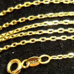 May include: Close-up of a gold necklace with a delicate chain design. The necklace features a small clasp and a tag with the marking "750". The chain is made of small, rectangular links.