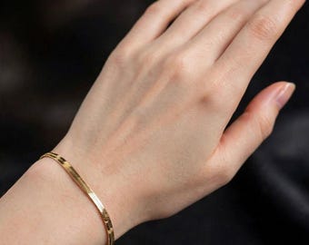 Può includere: Un bracciale a catena a spina di pesce color oro caratterizzato da un design a maglie piatte, flessibili e fittamente intrecciate che poggia sul polso. La superficie metallica presenta una finitura lucida e riflettente che cattura la luce.