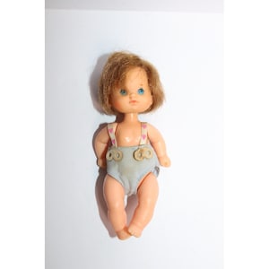 May include: A vintage doll with short, light brown hair and blue eyes. The doll is wearing a light blue nappy with wooden button details and pink heart-patterned braces. The doll is standing on a white surface.