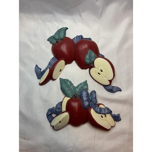 May include: Two sets of decorative apple wall hangings. Each set features a red apple, a sliced apple, and green leaves. Blue ribbons accent the apples. The background is a white fabric.