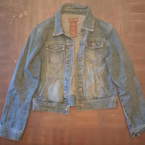 May include: A light blue denim jacket with a classic design. The jacket features a collared neckline, button-down closure, two chest pockets, and two side pockets. The jacket has a faded wash and a label that says "Jella's".