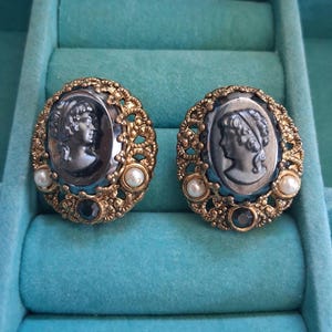 May include: A pair of oval cameo earrings. Each earring features a gray profile within a gold-toned filigree frame, accented with faux pearls and a dark blue stone. The earrings are displayed on a turquoise velvet surface.