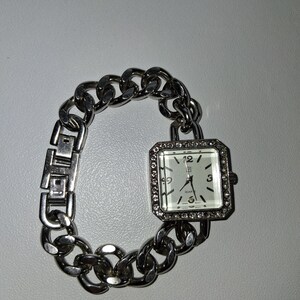 May include: A silver-toned wristwatch with a square face and a chain-link band. The watch face is white with black numerals and hands. The bezel is encrusted with small, clear crystals. The watch is designed to be worn on the wrist.