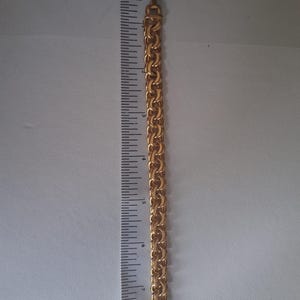 May include: A gold-colored chain bracelet with a textured, linked design. The bracelet is approximately 8 inches long. The clasp is visible at the end of the bracelet. The bracelet is next to a ruler.