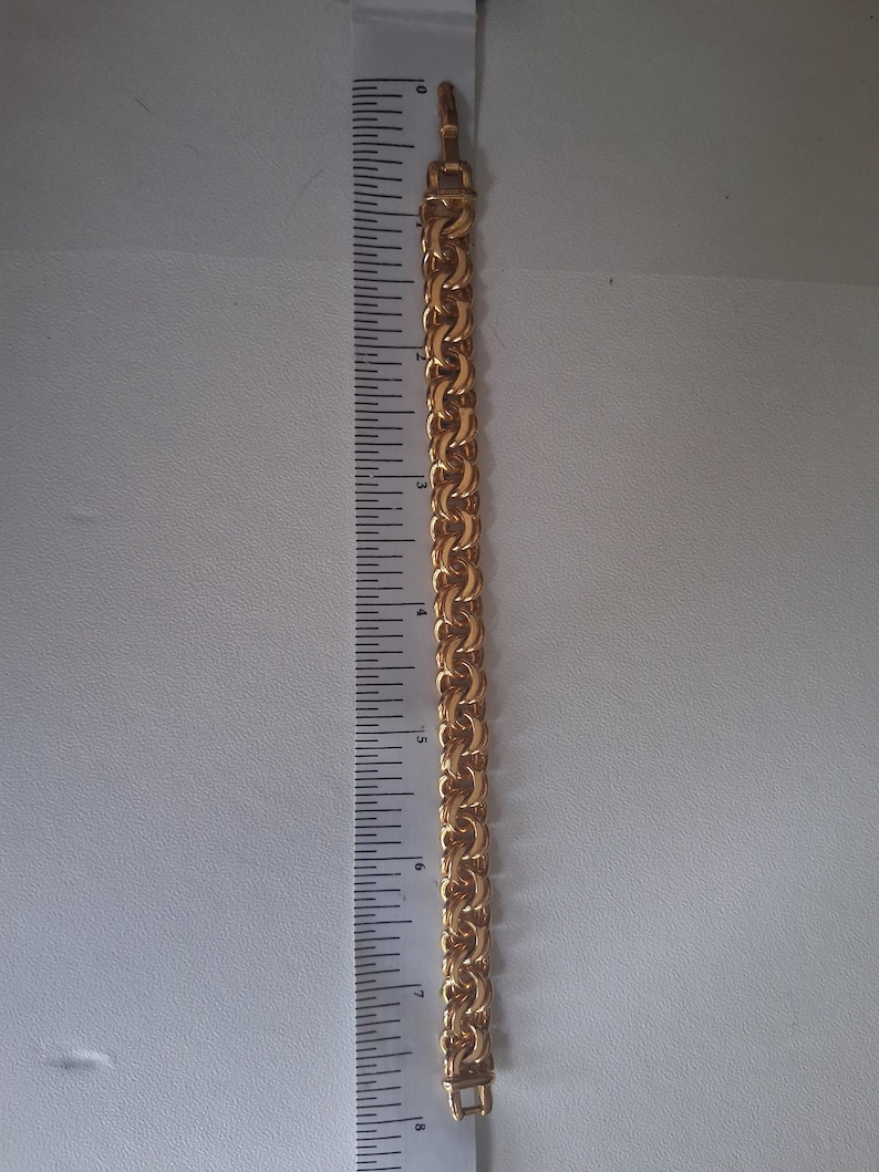 May include: A gold-colored chain bracelet with a textured, linked design. The bracelet is approximately 8 inches long. The clasp is visible at the end of the bracelet. The bracelet is next to a ruler.