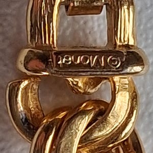 May include: Close-up of a gold-toned chain link bracelet. The bracelet features a series of interlocking, rounded links. The clasp is visible, with the brand name "Monet" stamped on it. The metal has a polished, reflective surface.