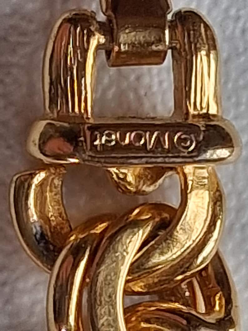May include: Close-up of a gold-toned chain link bracelet. The bracelet features a series of interlocking, rounded links. The clasp is visible, with the brand name "Monet" stamped on it. The metal has a polished, reflective surface.