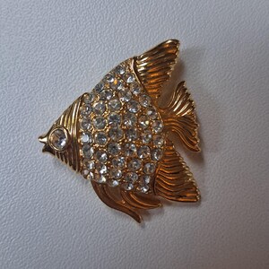 May include: A gold-toned fish-shaped brooch with clear, sparkling rhinestones covering the body. The brooch has detailed fins and a small eye, creating a realistic and elegant design. This jewelry piece is suitable for adding a touch of glamour to any outfit.