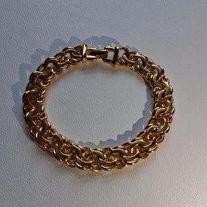 May include: A gold-colored bracelet with a thick, linked chain design. The bracelet is made of metal and features a secure clasp. The links are closely connected, creating a solid, circular shape. The bracelet is shown on a light gray surface.