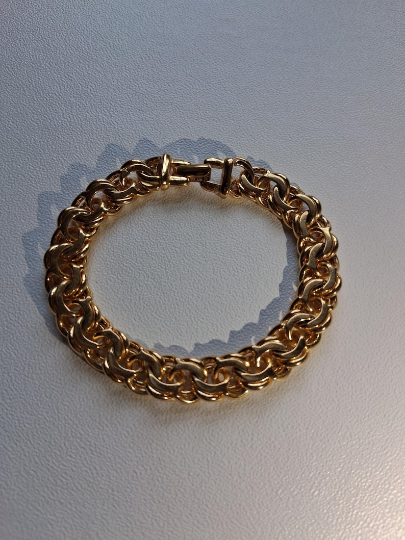 May include: A gold-colored bracelet with a thick, linked chain design. The bracelet is made of metal and features a secure clasp. The links are closely connected, creating a solid, circular shape. The bracelet is shown on a light gray surface.