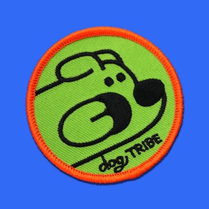 May include: Circular patch with a green background and an orange border. The patch features a black cartoon dog face, with the words "dog TRIBE" in black script below the dog.