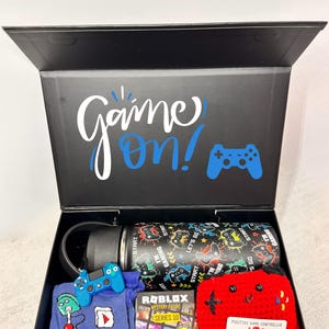 Puede incluir: Una caja de regalo negra con las palabras "Game On!" y un gr&aacute;fico de mando. En el interior hay una botella de agua negra con un dise&ntilde;o de juego, un llavero de mando azul, un su&eacute;ter azul, una figura misteriosa de Roblox y un mando de ganchillo rojo.