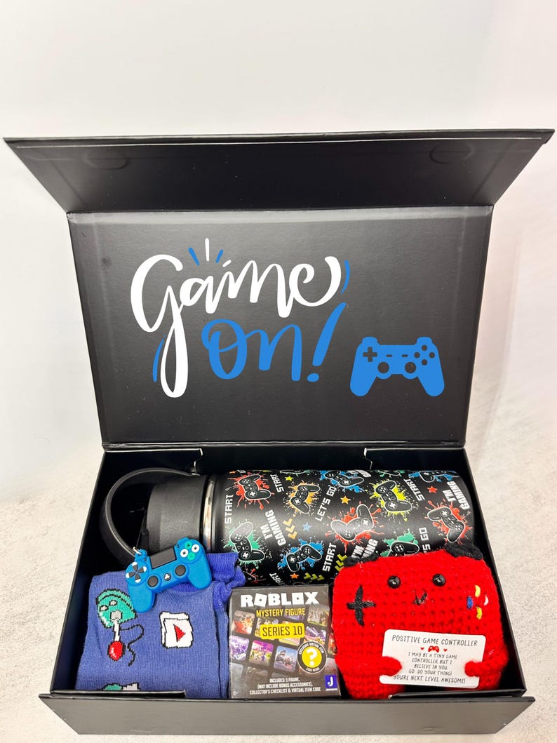 Puede incluir: Una caja de regalo negra con las palabras "Game On!" y un gr&aacute;fico de mando. En el interior hay una botella de agua negra con un dise&ntilde;o de juego, un llavero de mando azul, un su&eacute;ter azul, una figura misteriosa de Roblox y un mando de ganchillo rojo.