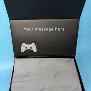 Puede incluir: Caja de regalo negra abierta con tapa abatible. El interior de la tapa tiene el texto "Your message here" y un icono blanco de mando de videojuegos. La caja contiene papel de seda gris con un logotipo.