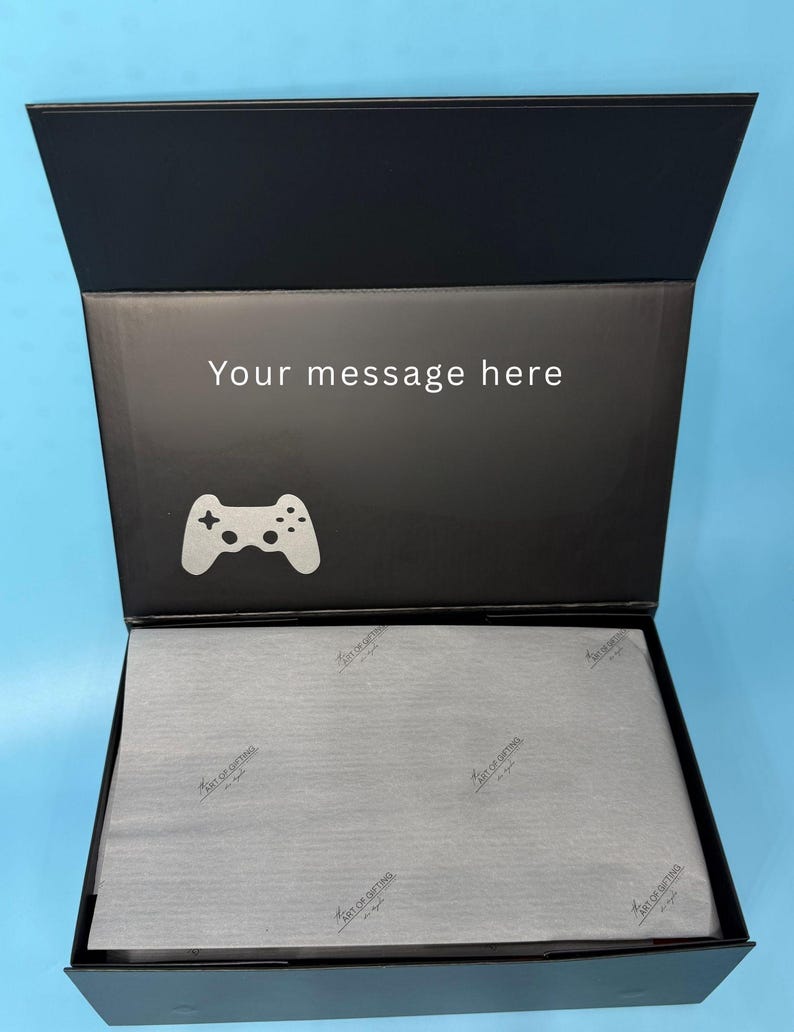 Puede incluir: Caja de regalo negra abierta con tapa abatible. El interior de la tapa tiene el texto "Your message here" y un icono blanco de mando de videojuegos. La caja contiene papel de seda gris con un logotipo.