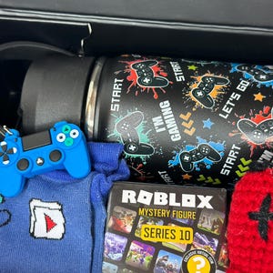 Puede incluir: Una caja de regalo negra que contiene art&iacute;culos con tem&aacute;tica de juegos. Incluye una botella de agua negra con gr&aacute;ficos de controlador, un llavero de controlador azul, un calcet&iacute;n azul con un dise&ntilde;o blanco y rojo, una caja de Roblox Mystery Figure y un art&iacute;culo de punto rojo.