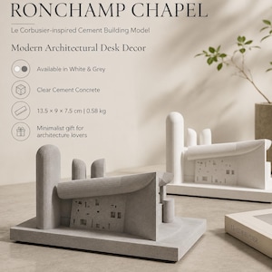 May include: Two cement building models of the Ronchamp Chapel, one in grey and one in white. Each model features a curved roof, a tall cylindrical tower, and a rectangular base. The dimensions are 13.5 x 9 x 7.5 cm with a weight of 0.58 kg.