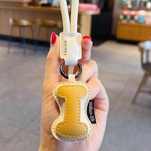 May include: A cream-coloured keychain with a yellow letter "I" charm. The charm is stitched onto a cream-coloured background. The keychain has a cream-coloured rope loop and a black metal ring. The brand name "YXDIAN" is printed on the leather strap.