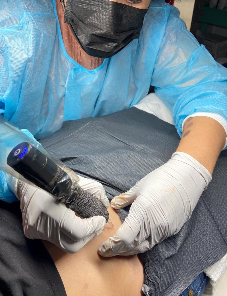 May include: A person wearing a blue protective gown and a black face mask is using a tattoo machine. The person is wearing white gloves and is working on a person's skin. The tattoo machine is black and has a blue light.