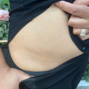 May include: Close-up of a person's midsection, wearing black clothing. The skin is light-toned, and the person's hand is visible, with a ring on the finger. The background includes greenery and a pink flower.