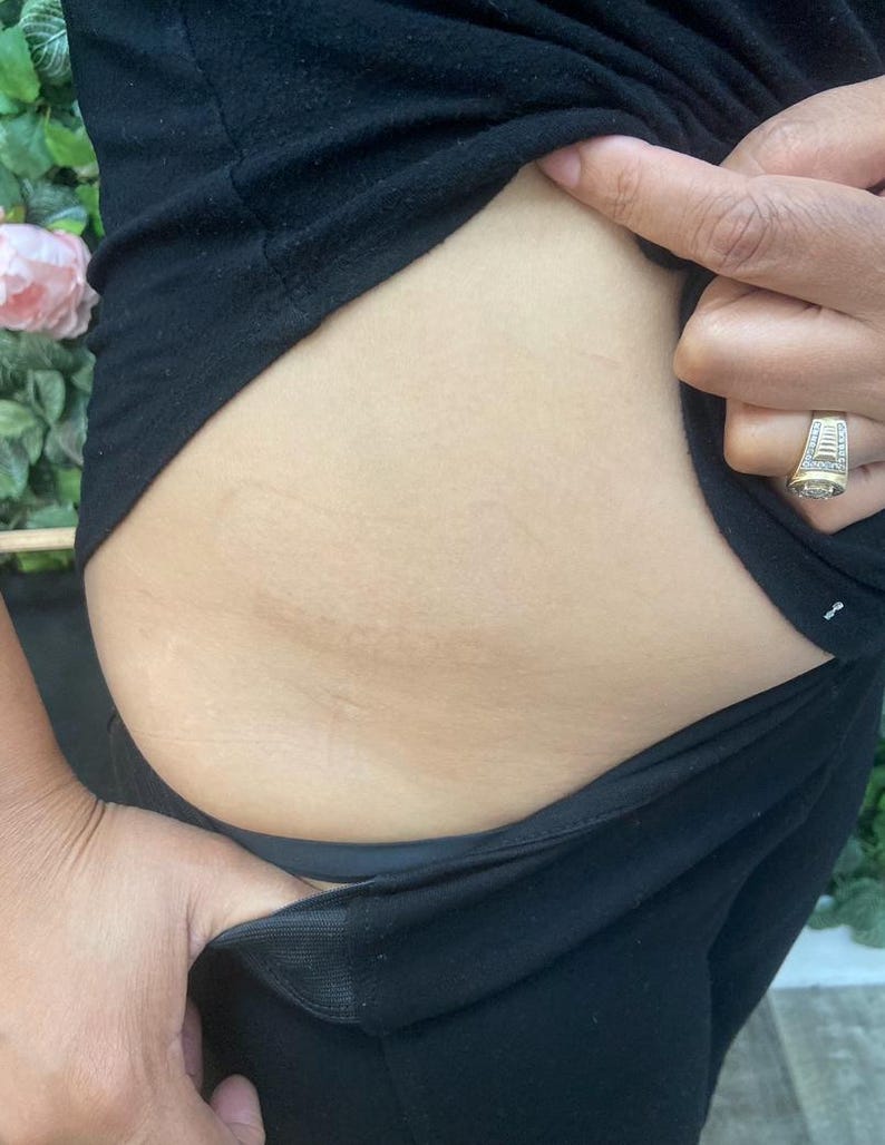 May include: Close-up of a person's midsection, wearing black clothing. The skin is light-toned, and the person's hand is visible, with a ring on the finger. The background includes greenery and a pink flower.