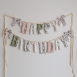 May include: A two-tiered banner with the words "HAPPY BIRTHDAY" in soft, pastel-colored fabric letters. The letters are adorned with floral patterns and bows, suspended by cream-colored ribbons, creating a festive decoration.