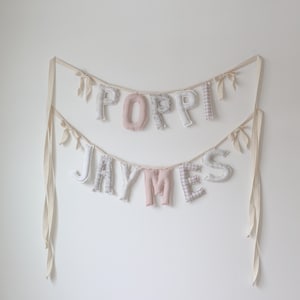 Op de afbeelding: Een dubbellaagse banner met de namen "POPPI" en "JAYMES" in stoffen letters. De letters zijn in wit, roze en patronen stoffen, opgehangen aan crèmekleurige linten met kleine strikjes. De banner hangt tegen een witte muur.