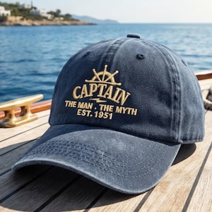 May include: A navy blue baseball cap with a distressed finish featuring gold embroidered text that reads CAPTAIN THE MAN THE MYTH EST. 1951, accompanied by a ship steering wheel icon centred above the word CAPTAIN on the front panel.
