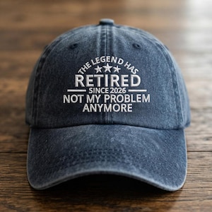 May include: A dark blue denim baseball cap with white embroidered text that reads THE LEGEND HAS RETIRED SINCE 2026 NOT MY PROBLEM ANYMORE, featuring three small stars above the word RETIRED and a curved brim with visible stitching details.