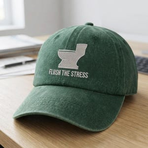 May include: A dark green baseball cap featuring an embroidered light grey graphic of a toilet and the text "FLUSH THE STRESS" stitched in white capital letters on the front panel. The cap has a curved brim and a structured crown design.