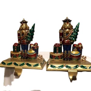 Puede incluir: Dos sujetadores de calcetines navideños con un soldado cascanueces sosteniendo un árbol de Navidad verde. Los sujetadores son dorados con detalles en rojo, azul y verde, y presentan cajas de regalo y adornos.