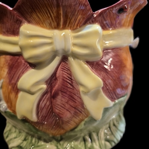 May include: A ceramic jug with a floral design, featuring a large yellow bow. The jug has a brown and burgundy colour scheme with green accents at the base, creating a charming, decorative piece.