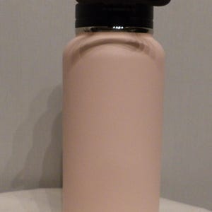 K&ouml;nnte beinhalten: Eine hellrosa isolierte Wasserflasche mit einem schwarzen Griff und Deckel. Die Flasche hat eine matte Oberfl&auml;che und ein kleines rundes Logo mit der Aufschrift "Life is Good". Die Flasche ist so konzipiert, dass Getr&auml;nke kalt oder hei&szlig; gehalten werden.