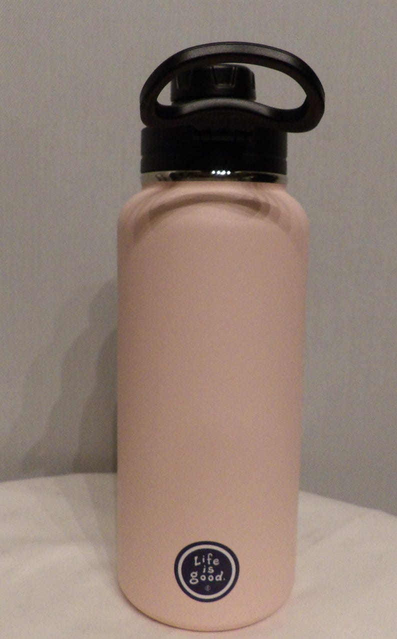 K&ouml;nnte beinhalten: Eine hellrosa isolierte Wasserflasche mit einem schwarzen Griff und Deckel. Die Flasche hat eine matte Oberfl&auml;che und ein kleines rundes Logo mit der Aufschrift "Life is Good". Die Flasche ist so konzipiert, dass Getr&auml;nke kalt oder hei&szlig; gehalten werden.