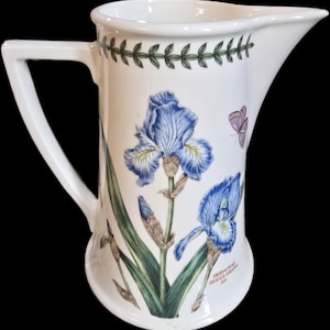 May include: A white ceramic pitcher featuring a botanical illustration of blue iris flowers, green leaves, and a small butterfly. The rim is decorated with a green leaf border, and the text IRIDACEAE DOUGLASIANA Iris is printed near the base.
