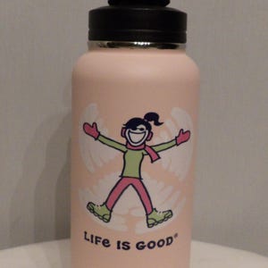 K&ouml;nnte beinhalten: Eine hellrosa isolierte Wasserflasche mit schwarzem Deckel. Die Flasche zeigt eine Grafik einer Person, die einen Schneeengel macht, mit gr&uuml;nem Hemd, rosa Hose und rotem Schal. Die Worte "Life is good" sind darunter gedruckt.