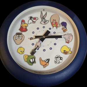 May include: A round wall clock with a dark blue frame and a white face featuring various Looney Tunes characters. The clock hands are shaped like an arm and a hand holding a mallet. The numbers one through twelve are printed in blue around the dial.